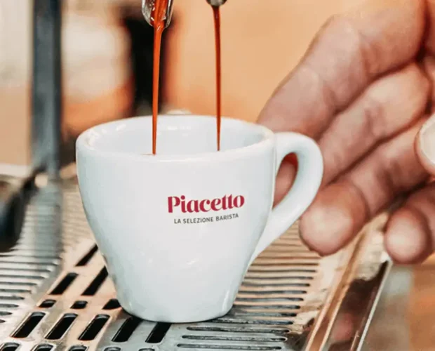 Χέρι ακουμπάει λευκό φλυτζάνι Piaccetto στο οποίο πέφτει φρεσκοκομμένος καφέ ςespresso από μηχανή espresso.