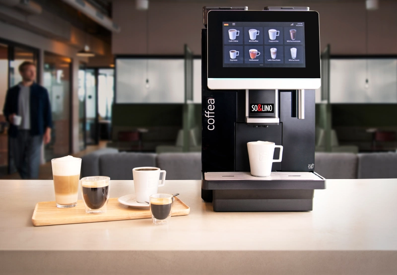 Η πλήρως αυτόματη μηχανή καφέ Solino coffea enjoy στον παγκο της κουζίνας ενός γραφείου με έναν φρεσκοπαρασκευασμένο καπουτσίνο και 4 ακόμη σπεσιαλιτέ καφέ στα αριστερά