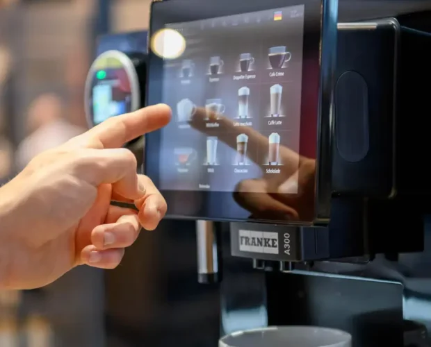Close-up of a person selecting a drink on a Franke A300 coffee machine's touchscreen, with a cup waiting below.