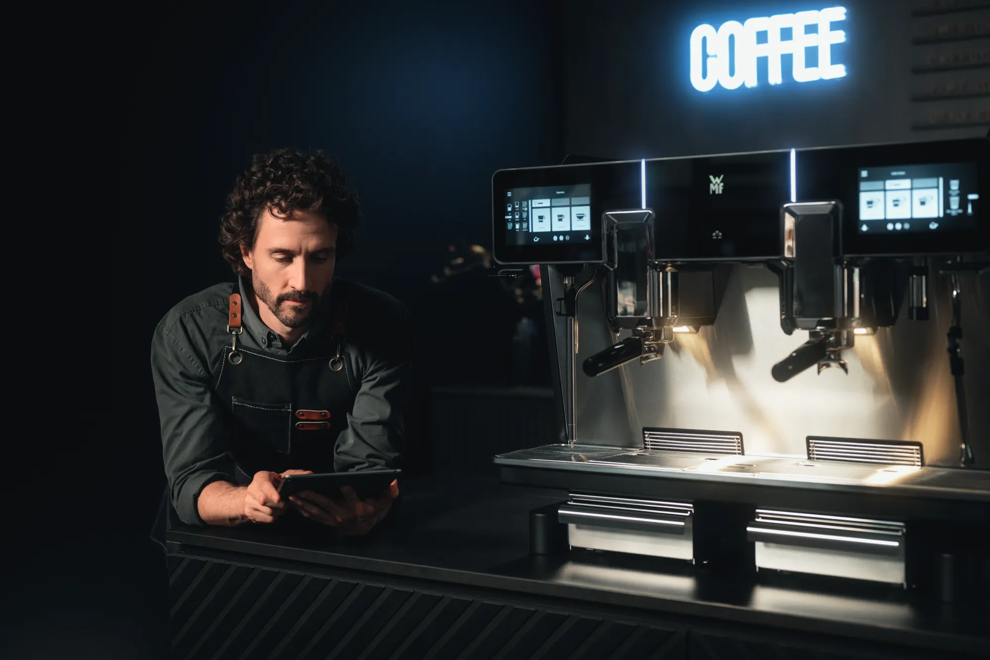 Focused barista in black apron using tablet next to a WMF espresso NEXT professional coffee machine, under neon 'COFFEE' sign, in a dark café setting.