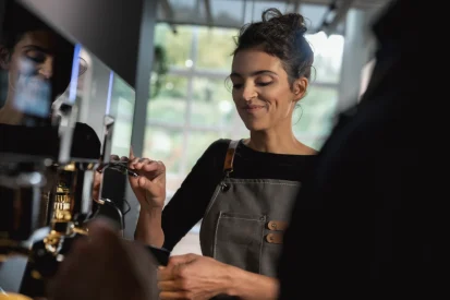 Barista preparing coffee with a WMF espresso NEXT coffee machine with a content smile, wearing a gray apron in a café setting with natural light.